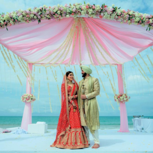 Bride and groom standing under a pink and floral decorated canopy by the beach.

